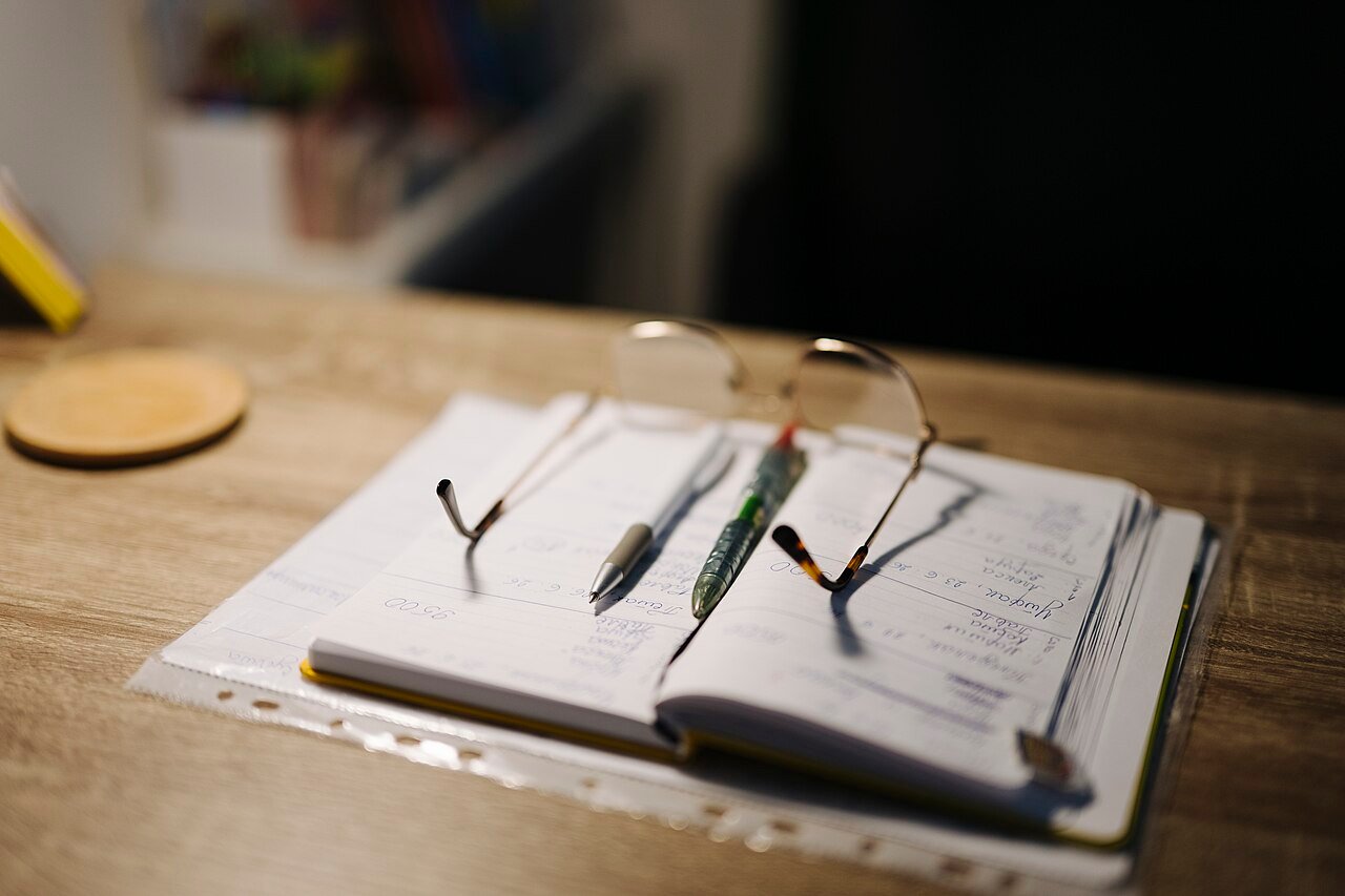 Cuaderno abierto con bolígrafos y gafas de lectura sobre una mesa